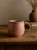 Różowy, teksturowany kubek ceramiczny stoi na rustykalnym drewnianym stole. W miękko rozmytym tle stosik książek oraz mały, terakotowy wazon z suszoną trawą dopełniają ciepłą, przytulną aranżację.